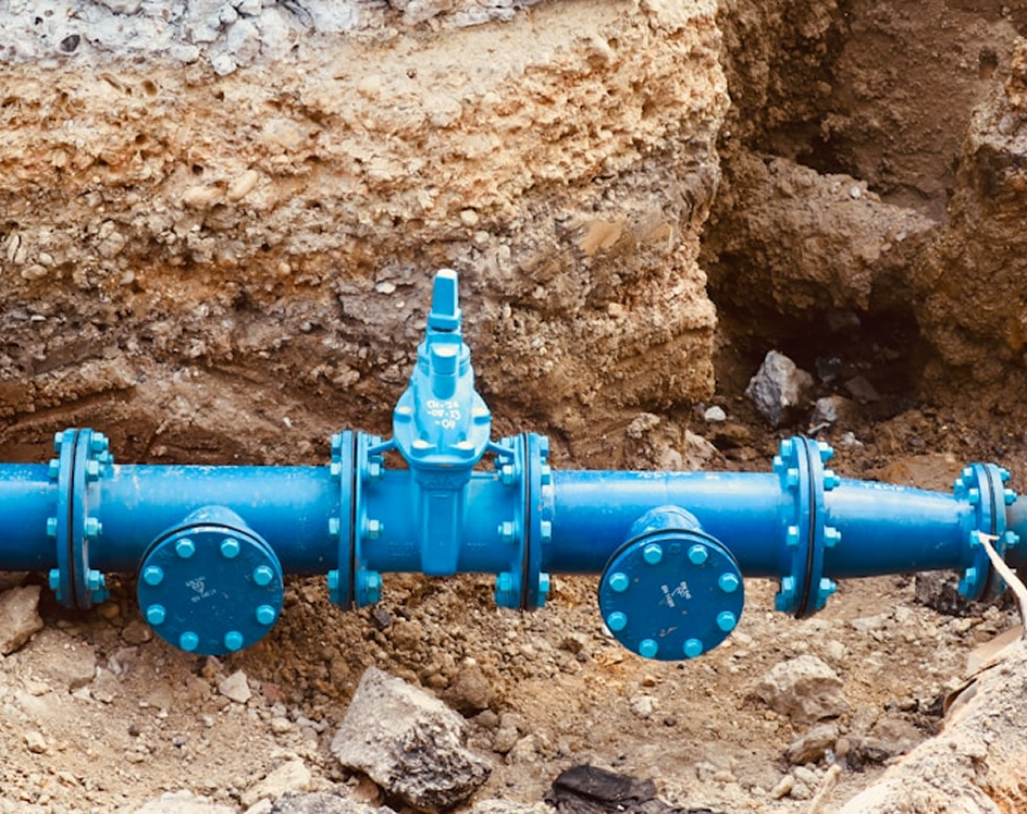 Blue underground water pipe with valves and flanges in a dirt trench.