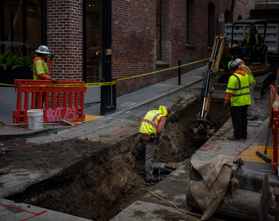 Construction workers in safety gear working on a deep trench on a city street beside brick buildings.